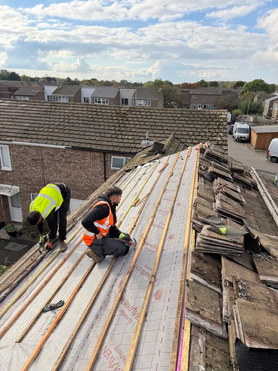 Two roofers fixing new battens over breathable membrane next to the stripped old tiles